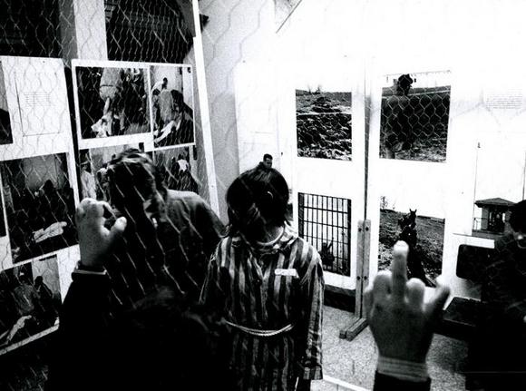 Uno spettatore guarda l'internata in isolamento, dietro la guardia e al muro fotografie di ospedali psichiatrici